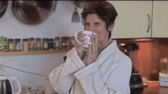 a woman in a bathrobe is drinking from a cup with a red flower on it