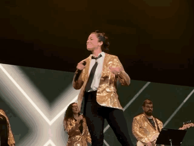 a woman in a gold jacket and tie is singing into a microphone on stage