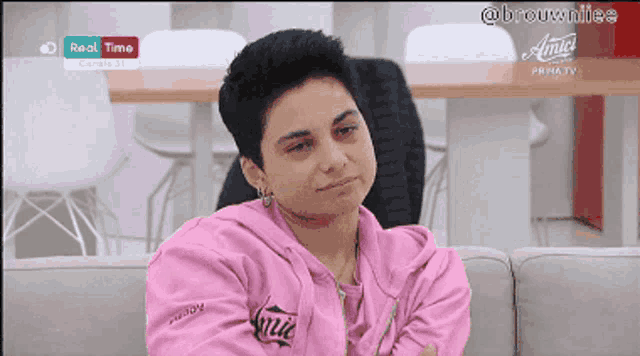 a woman in a pink sweatshirt is sitting on a couch in front of a sign that says real time