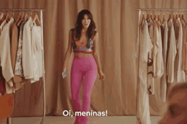 a woman in pink pants is standing in front of a rack of clothes and says " oi meninas "