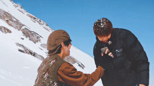 two men standing on top of a snow covered mountain one is holding a cell phone