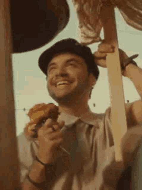 a man in a hat is holding a piece of wood and smiling while eating a pizza .