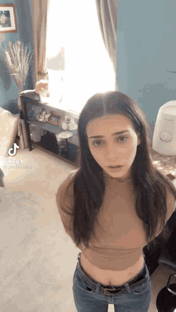 a woman is standing in a living room wearing a brown crop top and jeans .