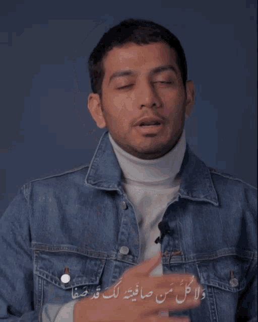 a man wearing a white turtleneck and a denim jacket has arabic writing on his chest