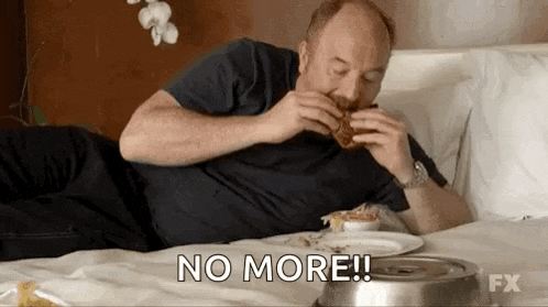 a man is laying on a bed eating a hamburger and says `` no more '' .