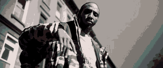 a black and white photo of a man wearing a camouflage jacket standing in front of a building .