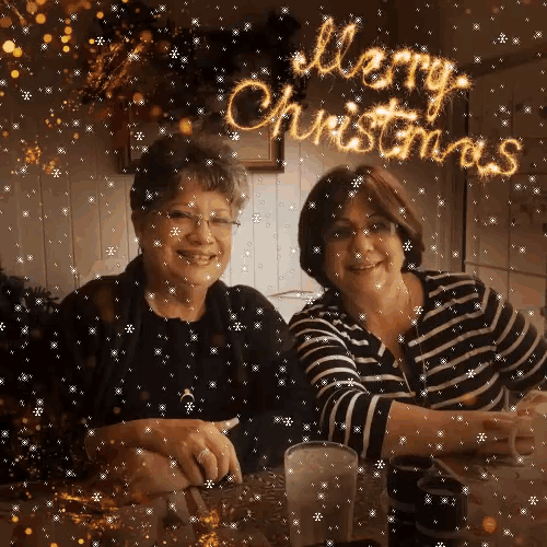 two women sitting at a table with a merry christmas sign behind them