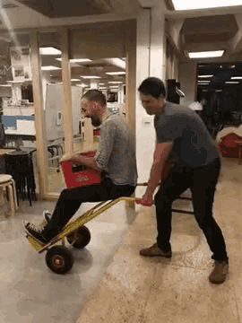 a man is pushing another man on a cart with a box on it .