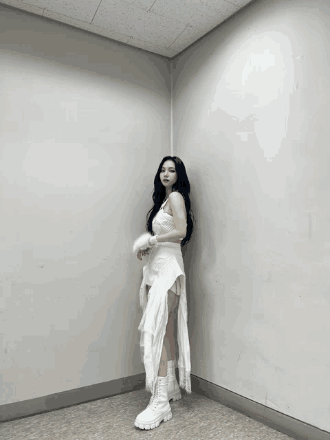 a woman in a white dress and boots stands in a corner of a room