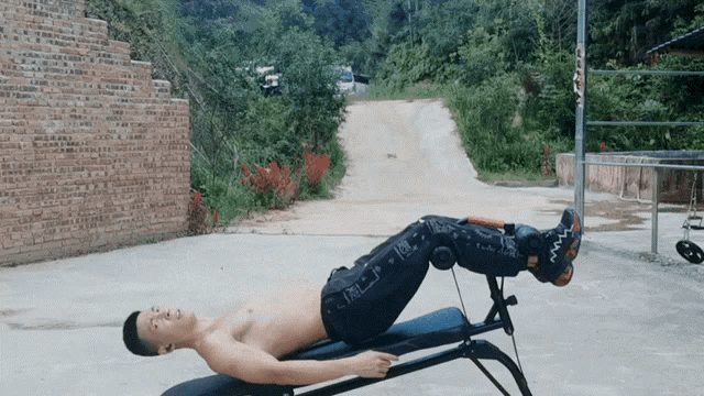 a shirtless man is laying on a bench with his legs crossed and a brick wall in the background