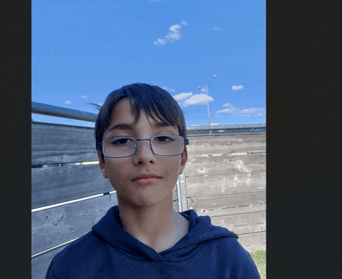 a boy wearing glasses and a black hoodie stands in front of a wooden fence