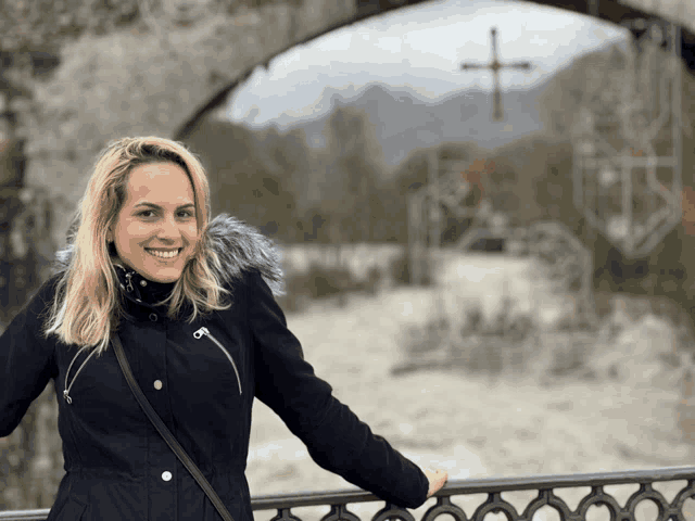 a woman in a black jacket with a fur hood is smiling in front of a cross