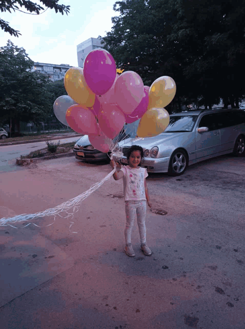 a little girl holding a bunch of pink and yellow balloons in a parking lot
