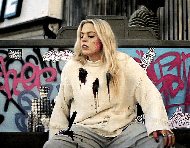 a woman wearing a white sweater is sitting in front of a wall that has graffiti on it including the letters rrp