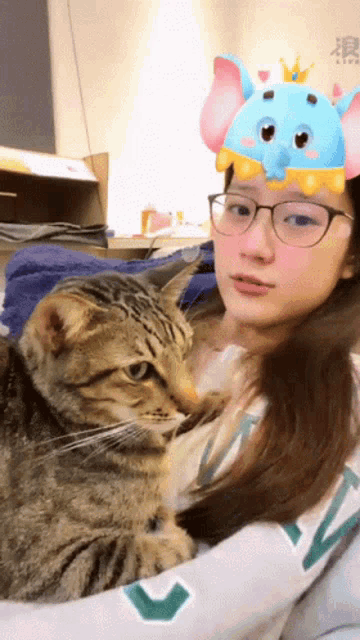 a woman wearing glasses holds a cat with an elephant headband on