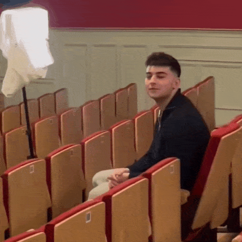 a young man sits in a row of wooden seats in a theatre