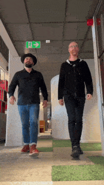 two men walk down a hallway with a green exit sign on the ceiling