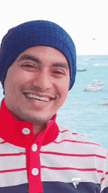 a man wearing a blue hat and a striped shirt smiles in front of the ocean