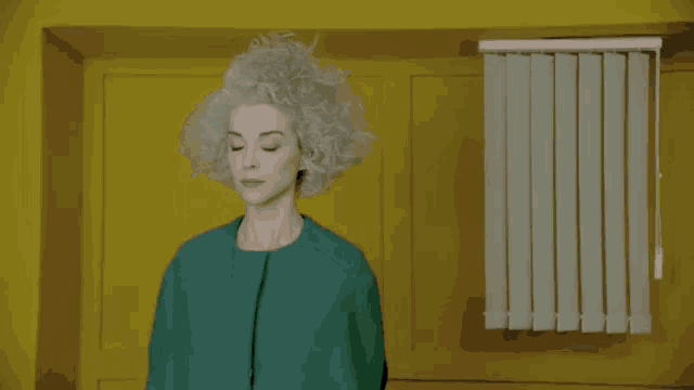 a woman with gray hair and a green coat is standing in front of a yellow wall and blinds .
