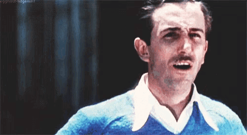 a man with a mustache is wearing a blue sweater and white shirt and making a funny face .