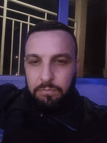 a man with a beard looks at the camera in front of a blue window