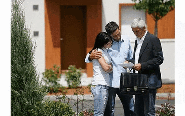 a man in a suit is holding a briefcase and talking to a couple standing in front of a house .
