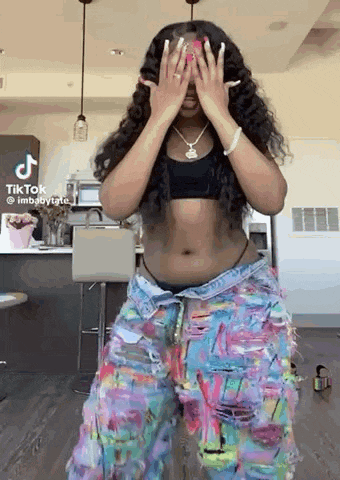 a woman is dancing in a kitchen wearing a crop top and colorful jeans .