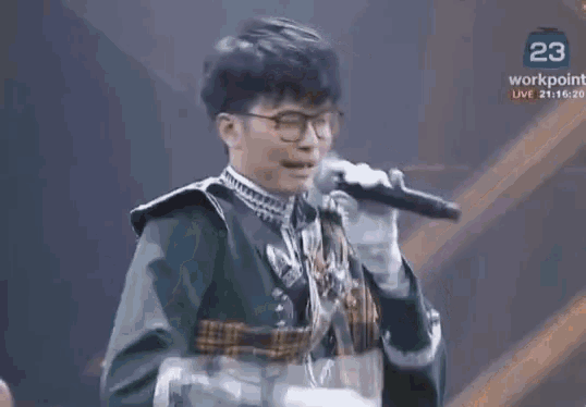 a man is singing into a microphone while wearing glasses and a green jacket .