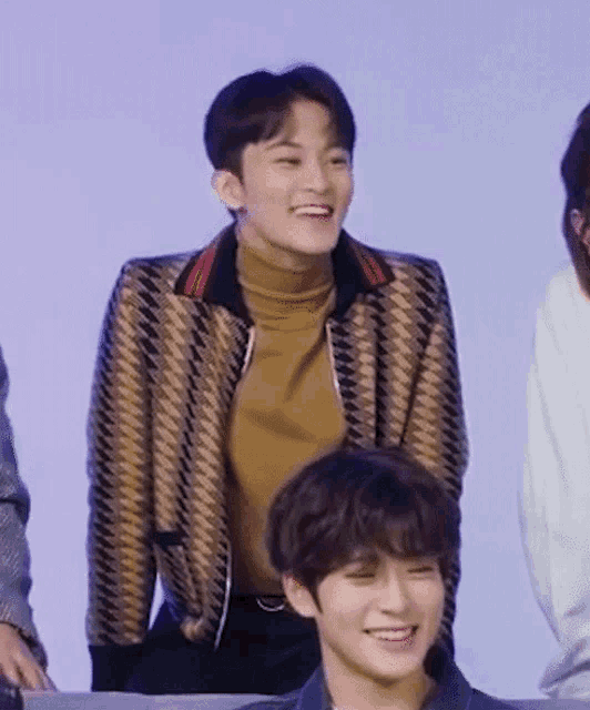 a man wearing a jacket and a turtleneck is smiling while sitting next to another man .