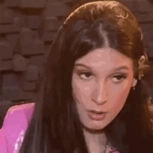 a woman with long dark hair is wearing a pink jacket and making a funny face .