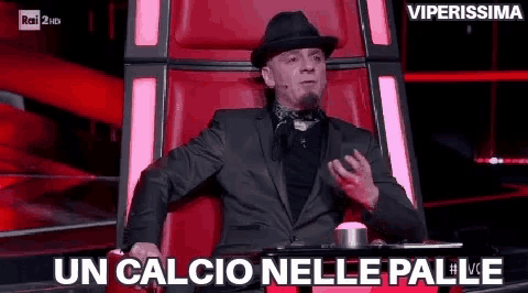 a man in a suit and hat is sitting in a chair that says un calcio nelle palle .