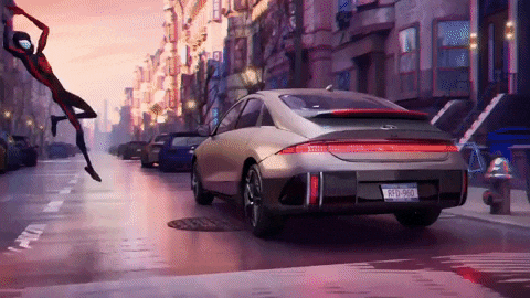 a silver kia car is driving down a city street with a spider-man flying in the background