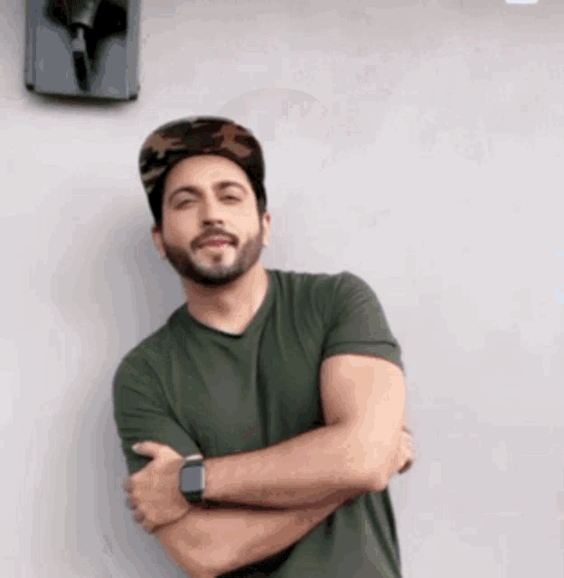 a man with a beard wearing a hat and a watch stands with his arms crossed .