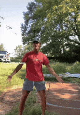 a man in a red shirt and shorts is dancing in a field with trees in the background