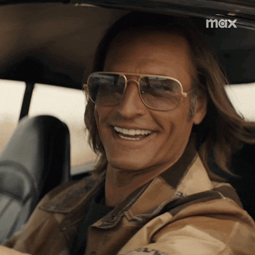 a man wearing sunglasses is smiling while sitting in a car with a max logo in the background