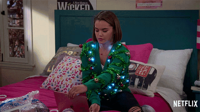 a girl in a green sweater sits on a bed using a laptop with netflix written on the bottom right