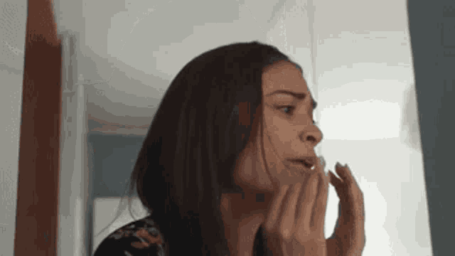 a woman is looking at her face in the mirror and covering her mouth with her hands .
