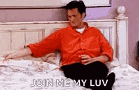 a man in a red shirt is sitting on a bed with a drink in his hand and says `` join me my luv '' .