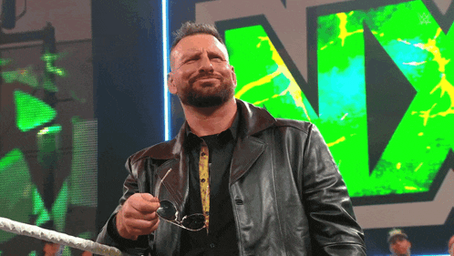 a man in a leather jacket holds a pair of sunglasses in front of a large green nx logo