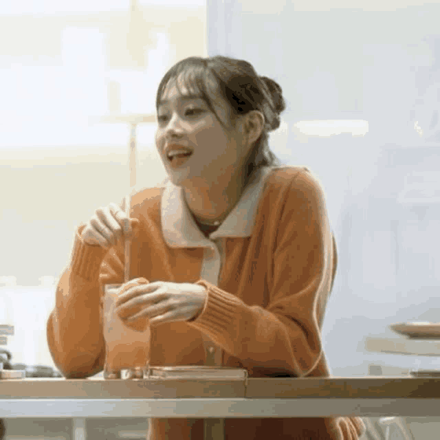 a woman in an orange sweater is sitting at a table drinking from a glass .