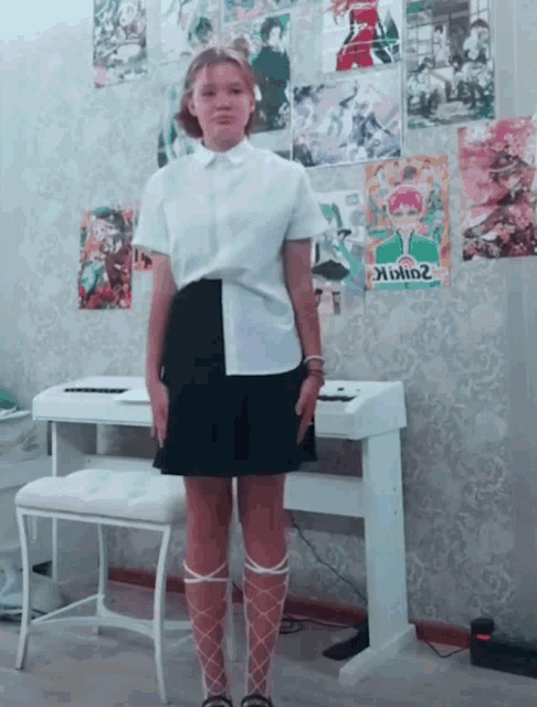 a girl in a white shirt and black skirt stands in front of a piano with posters on the wall including one that says goblin