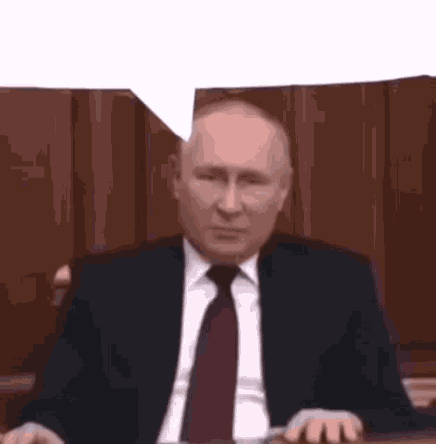 a man in a suit and tie is sitting at a desk with a speech bubble .