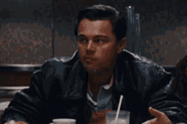 a man in a leather jacket is sitting at a table talking to another man .