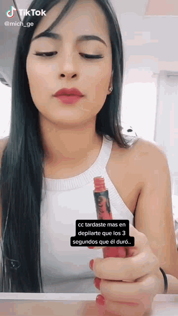 a woman in a white tank top is holding a red lip gloss in her hand