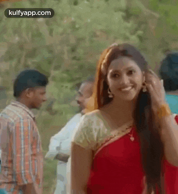 a woman in a red saree is smiling and looking at the camera while a man stands behind her .