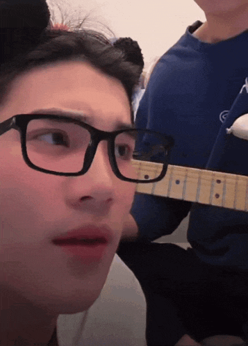 a man wearing glasses looks at the camera while another man holds a guitar