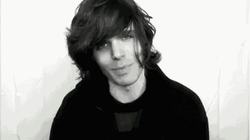a black and white photo of a young man with long hair smiling and looking at the camera .