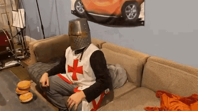 a man in a crusader costume is sitting on a couch eating a hamburger .