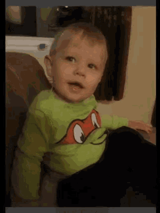 a baby wearing a green turtle shirt is sitting on a couch .