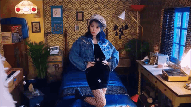 a woman with blue hair and a denim jacket is kneeling on a bed in a room .
